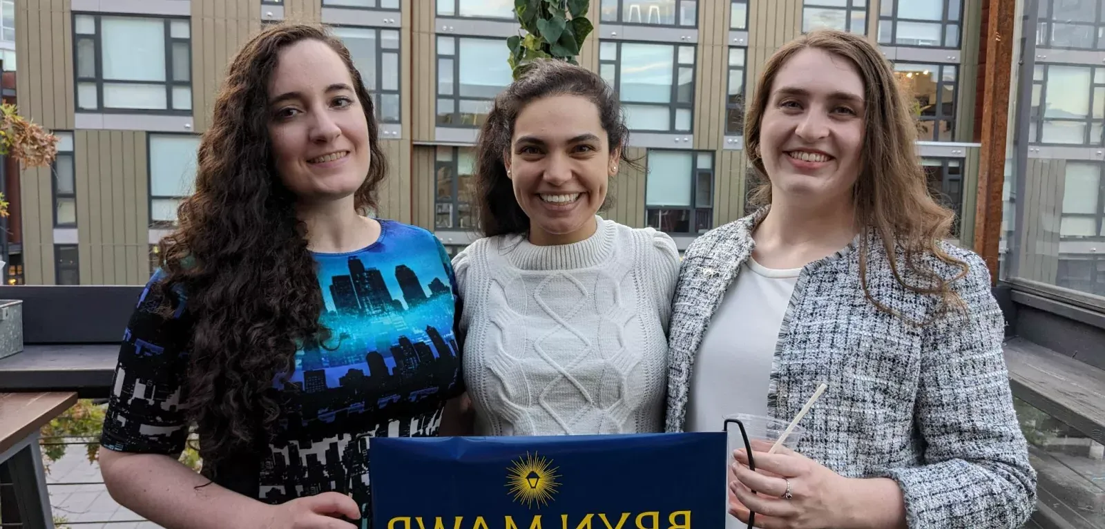 Three young alums smile at the camera. The are on the balcony of a building.